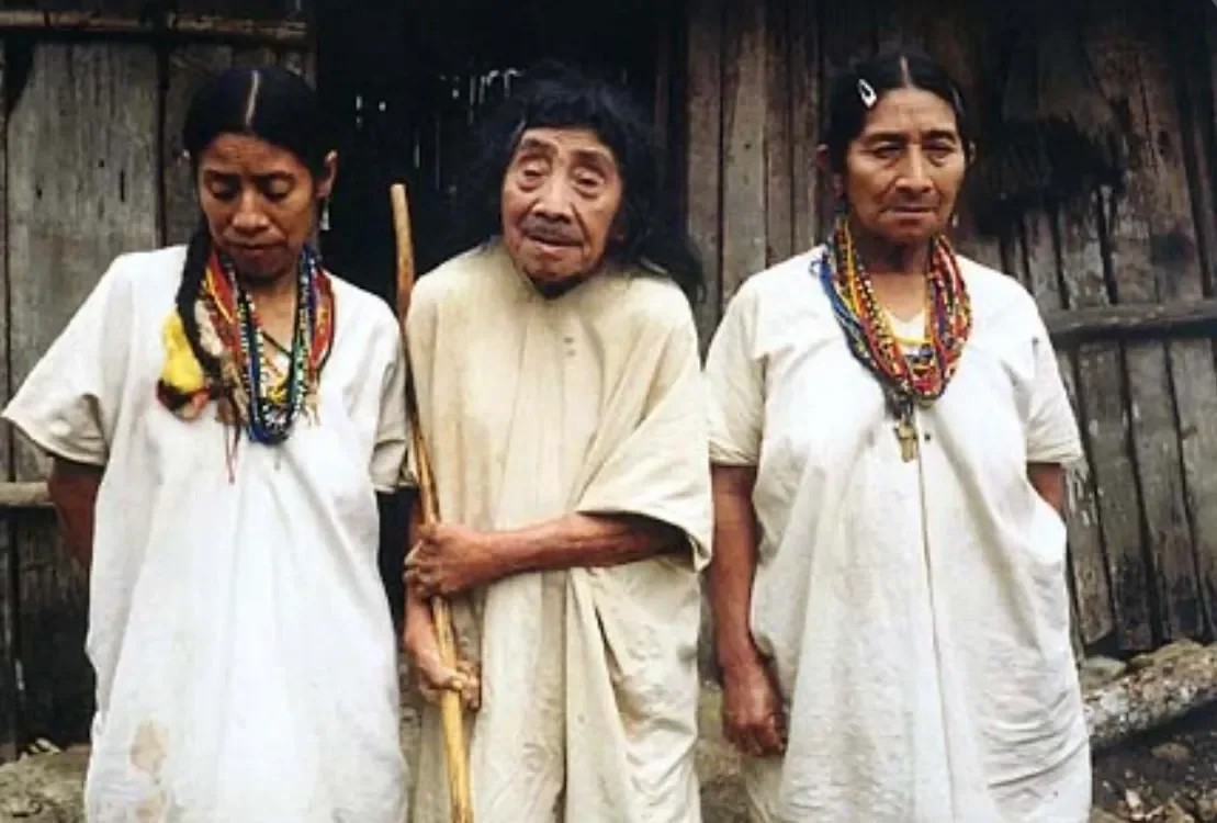 Three Lacandón Maya women in white tunics outside a wooden house